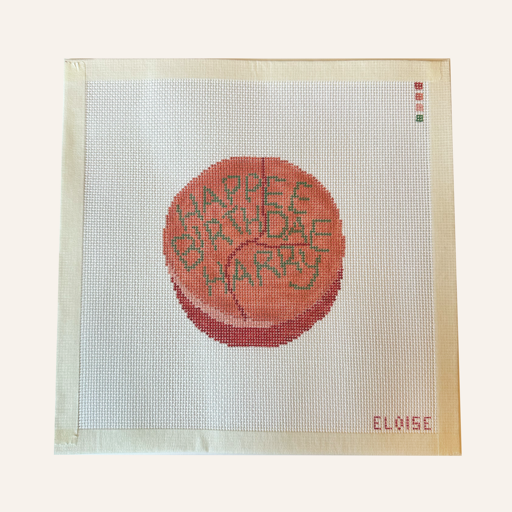 ​​A needlepoint canvas featuring a pink cake reading "Happee Birthdae Harry". The canvas features a color palette guide and is signed "Eloise" 