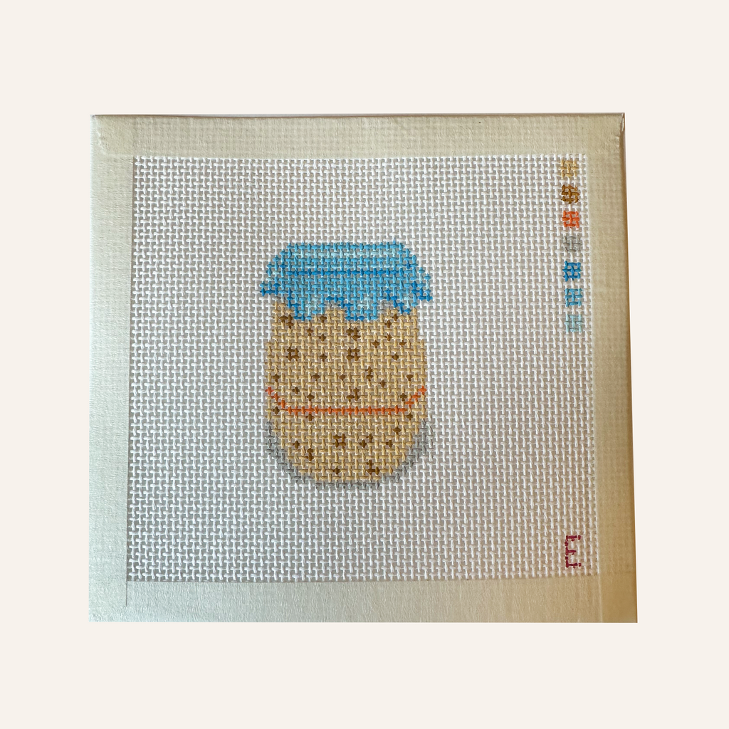 A needlepoint canvas featuring a sourdough starter pattern. The pattern showcases a jar filled with tan sourdough starter, an orange rubber band and blue  lid cover. The canvas features a color palette panel and is signed by "Eloise."