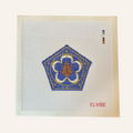 A chocolate frog needlepoint canvas designed with a blue pentagon shape with gold ornate detailing.  brown frog sits in the middle. the canvas has a color palette guide and is signed "Eloise"